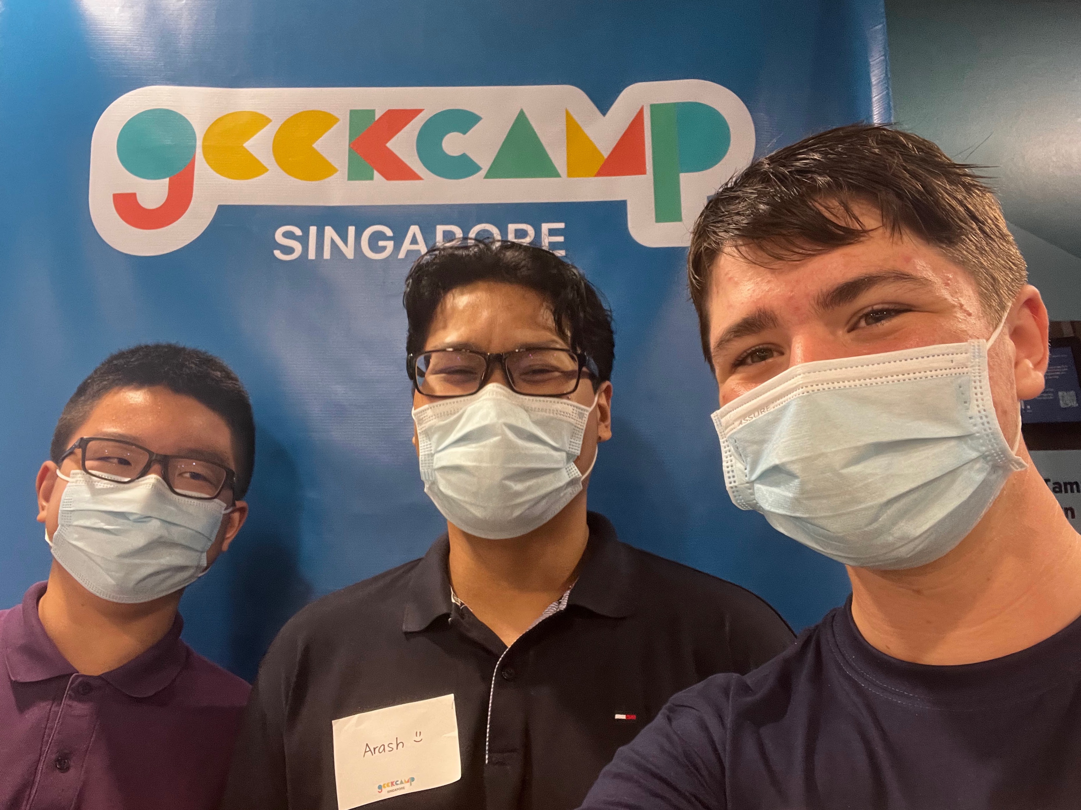 Three people smiling in front of a vertical banner with the words 'Geekcamp Singapore' on it